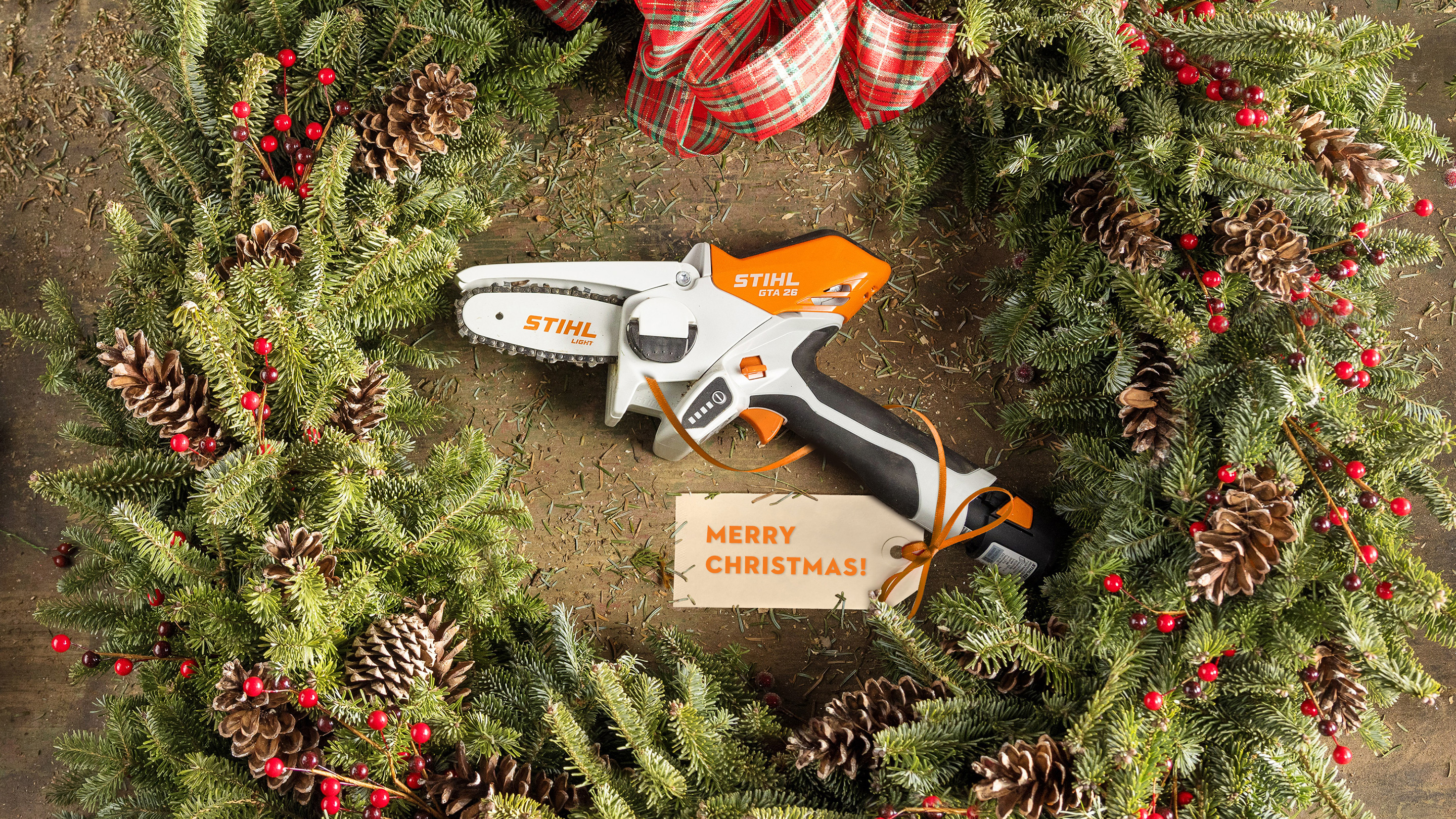 STIHL GTA 26 σε ένα χριστουγεννιάτικο στεφάνι, περιτριγυρισμένο από πράσινο έλατο, κουκουνάρια και κορδέλα με χριστουγεννιάτικη κάρτα